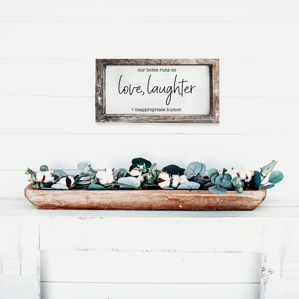 Decorative sign with text above a wooden tray with plants on a white surface.
