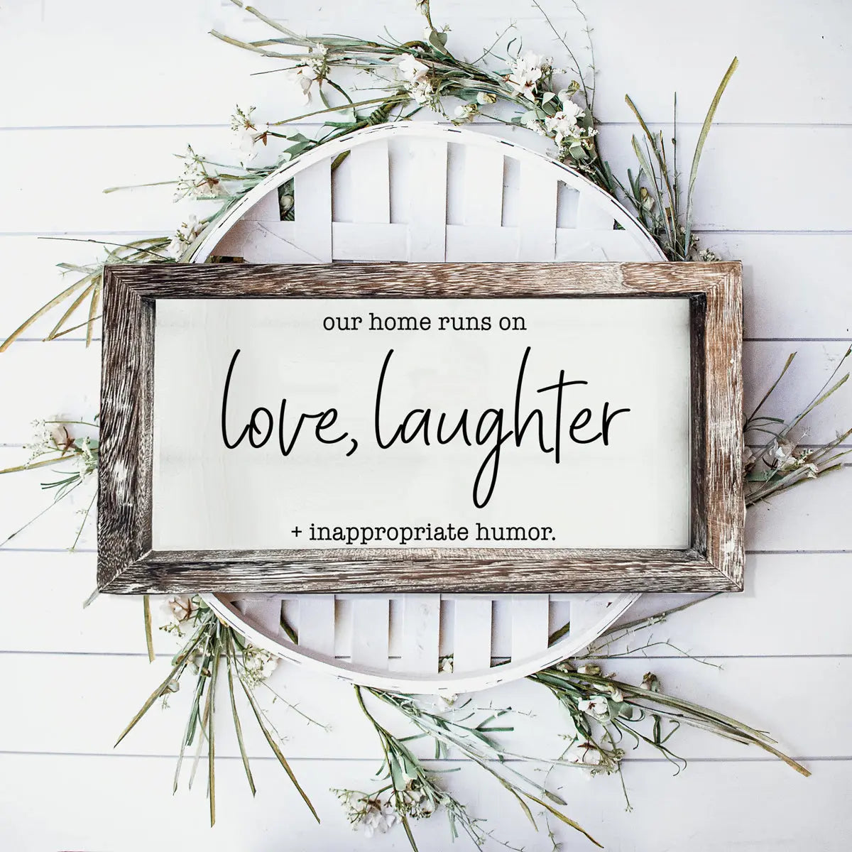 Wooden-framed sign with text 'our home runs on love, laughter + inappropriate humor' on a white background with greenery.