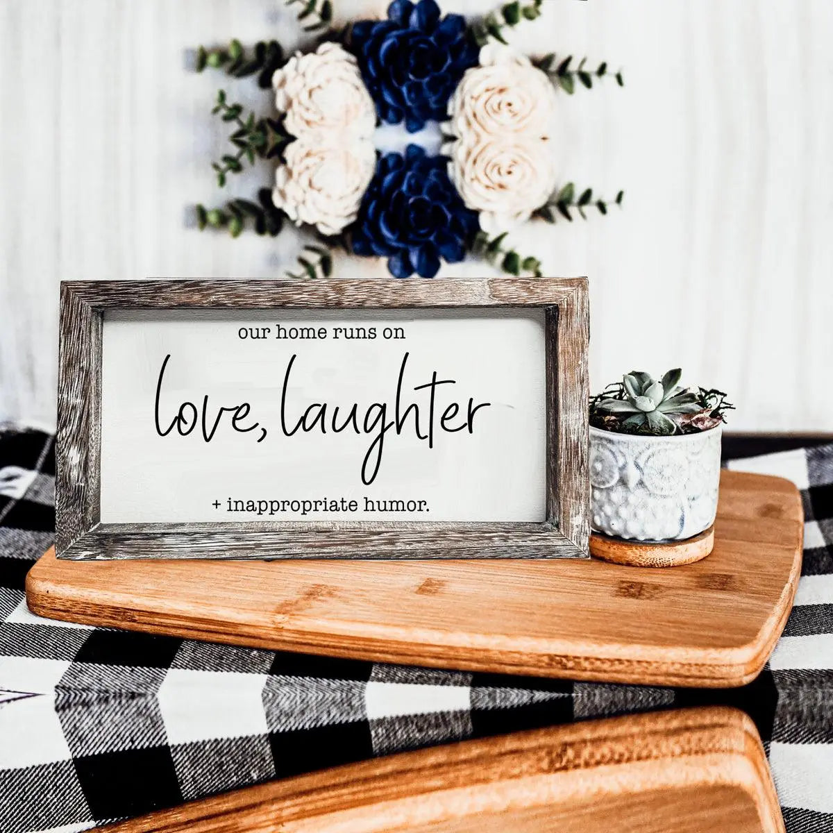 Wooden sign with 'love, laughter + inappropriate humor' text on a table with flowers in the background