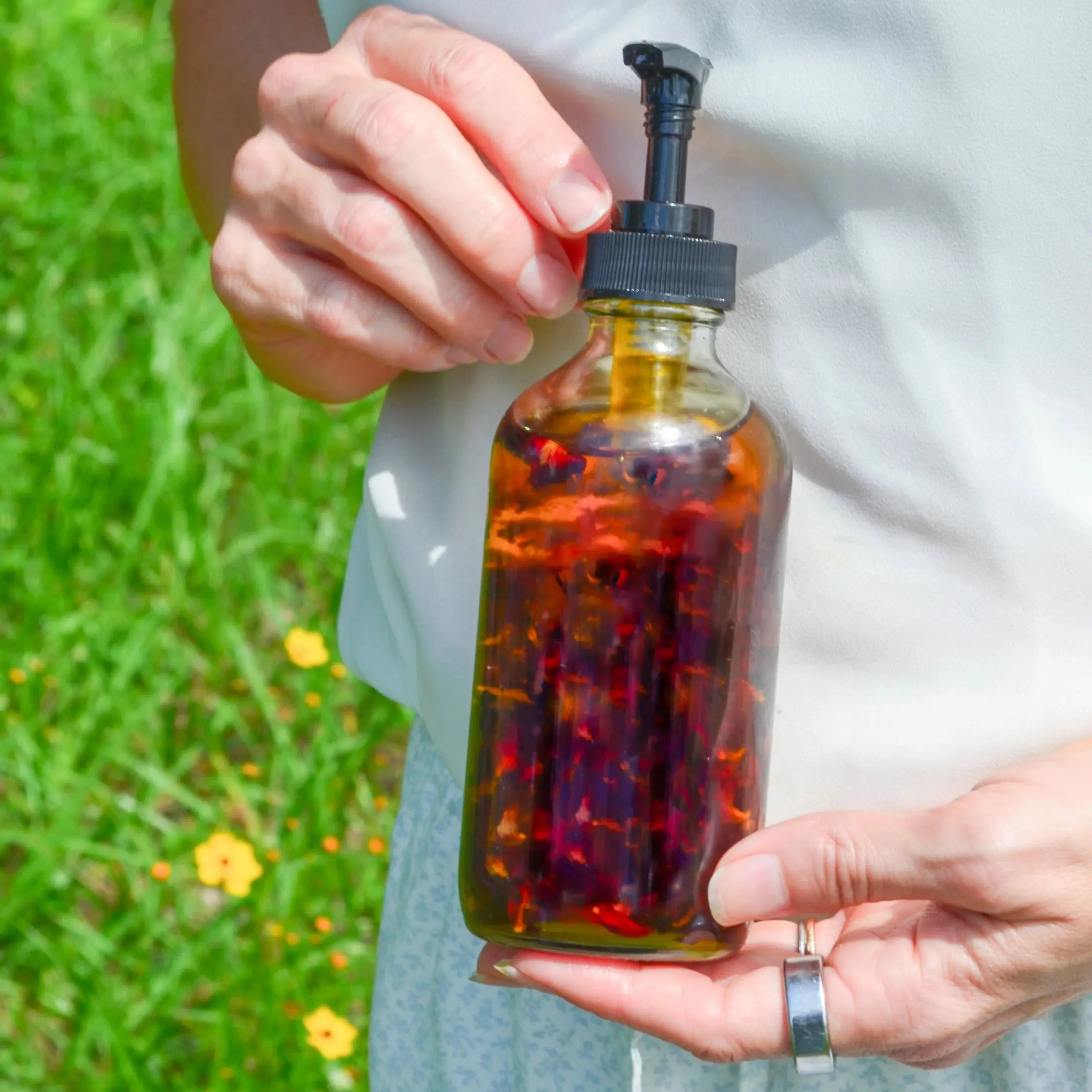 Hand holding glass bottle with floral bath oil, black pump, outdoor green grass background