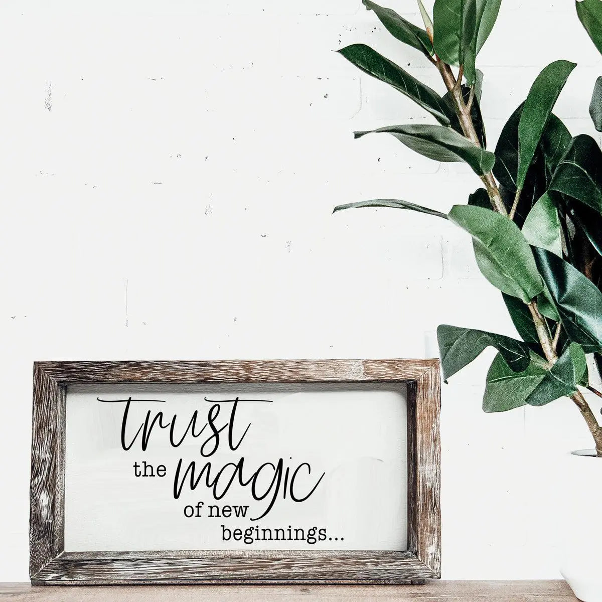 Framed sign with 'Trust the magic of new beginnings' text on a white wall with a plant.