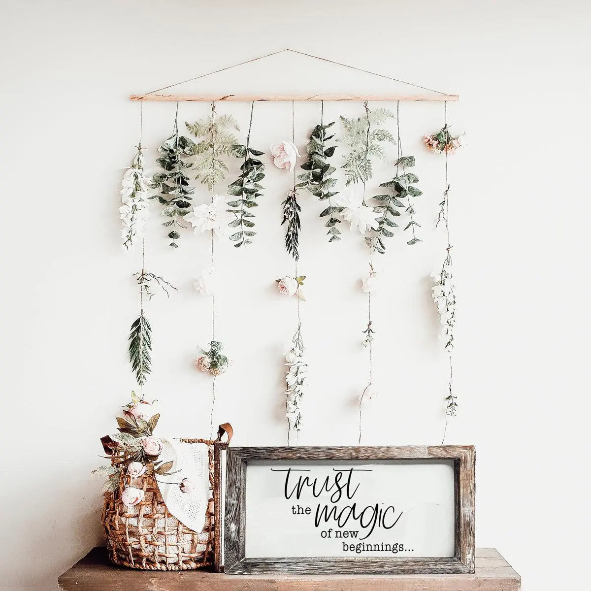 Decorative wall hanging with plants and a framed sign on a white wall.