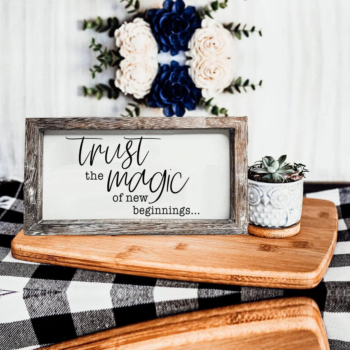Decorative sign with 'Trust the magic of new beginnings' text on a wooden surface with flowers in the background.