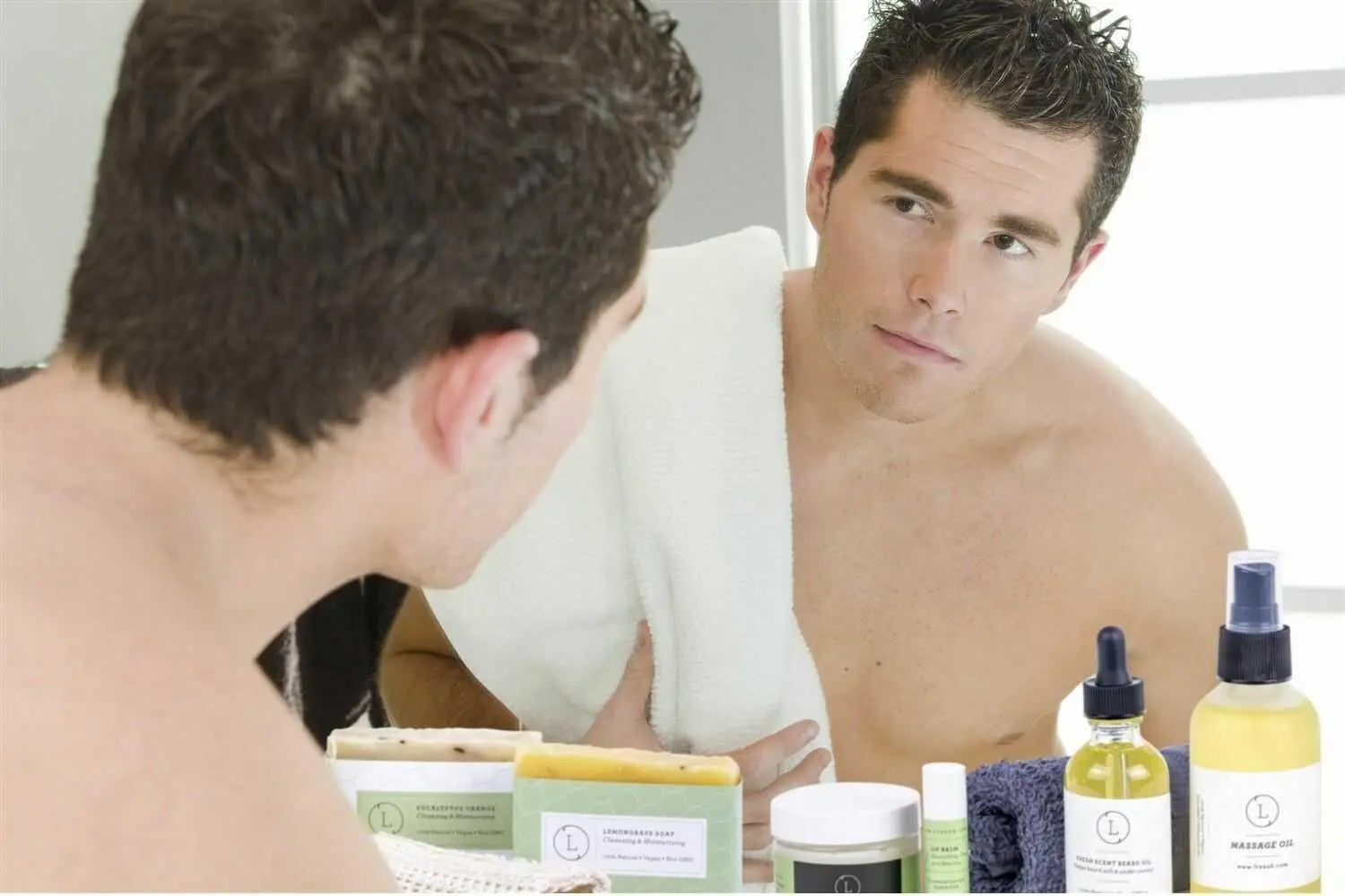 Man checking beard in mirror with natural bath essentials, soap, and massage oil for men's grooming