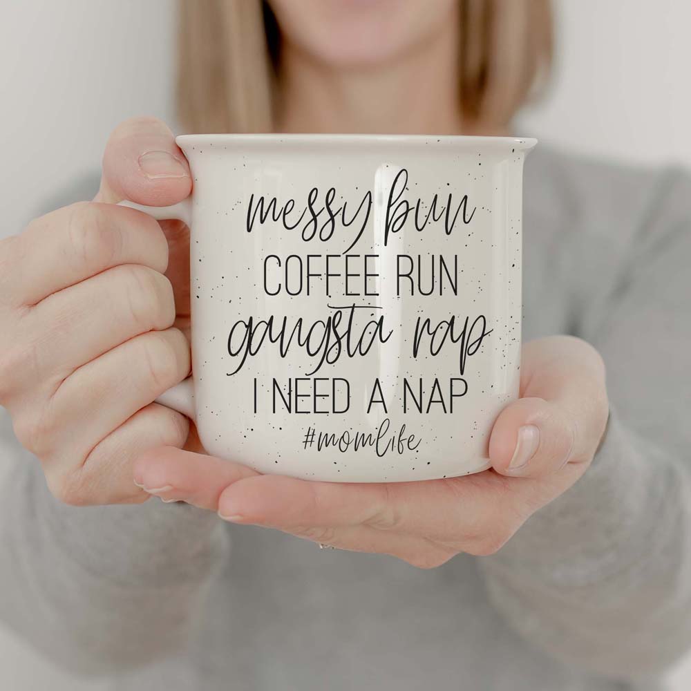 Person holding a mug with text 'messy bun coffee run gangsta rap I need a nap #momlife' against a neutral background