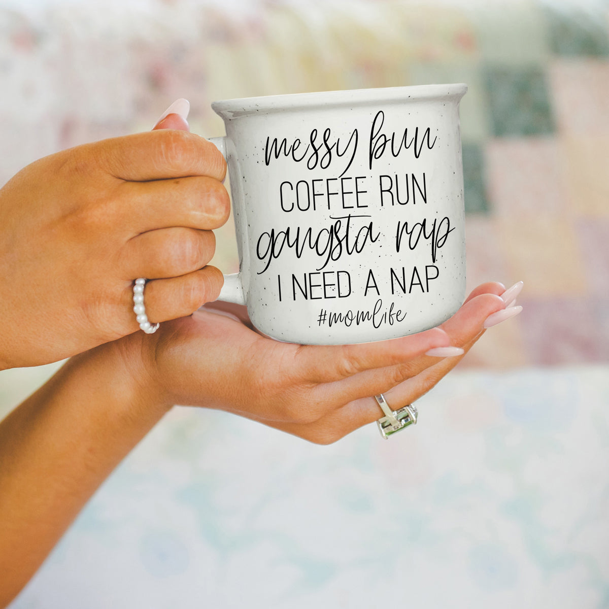 Hand holding a mug with text 'messy bun coffee run gangsta rap I need a nap #momlife' against a blurred background.