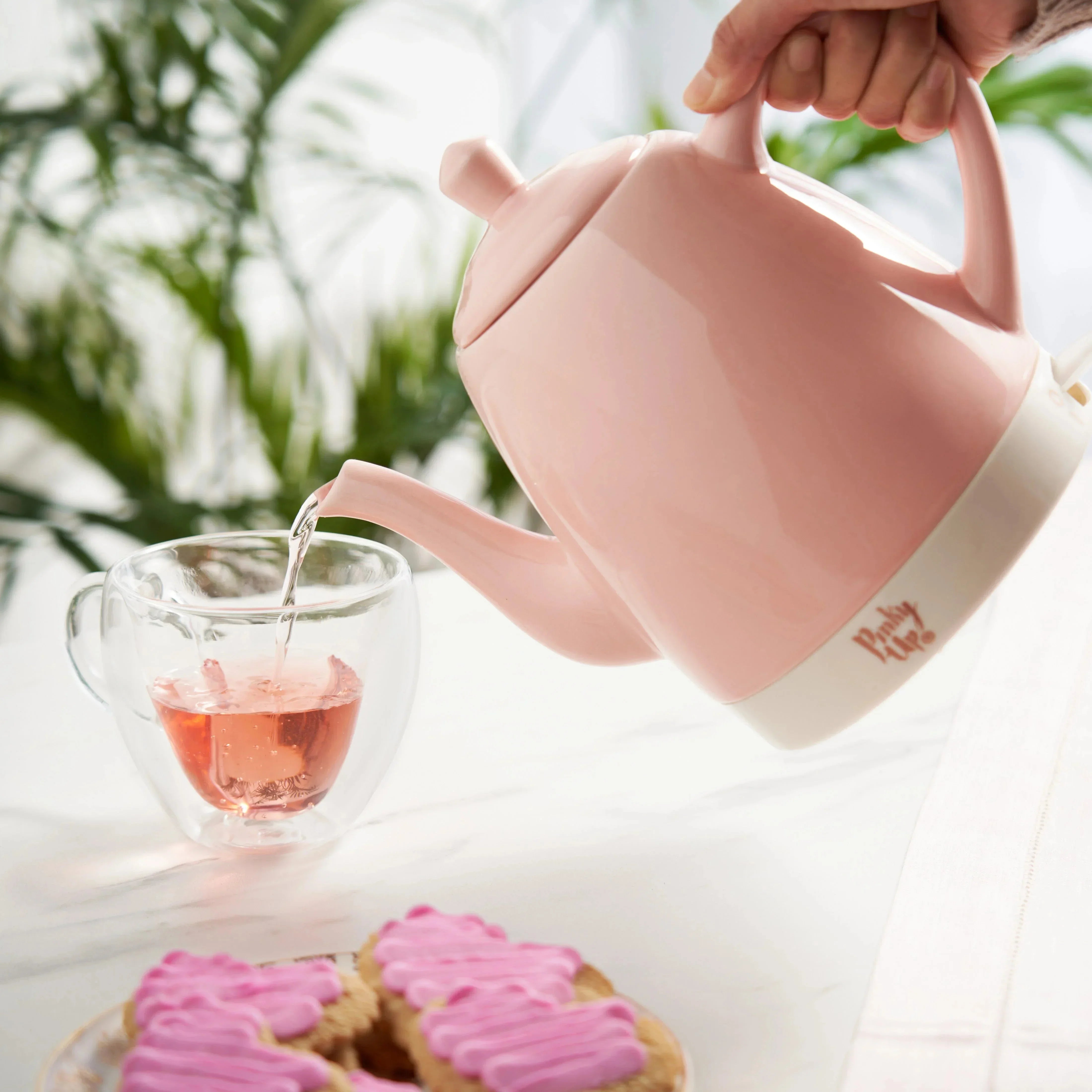 Pink teapot pouring tea into a glass cup with pink cookies on a white surface.