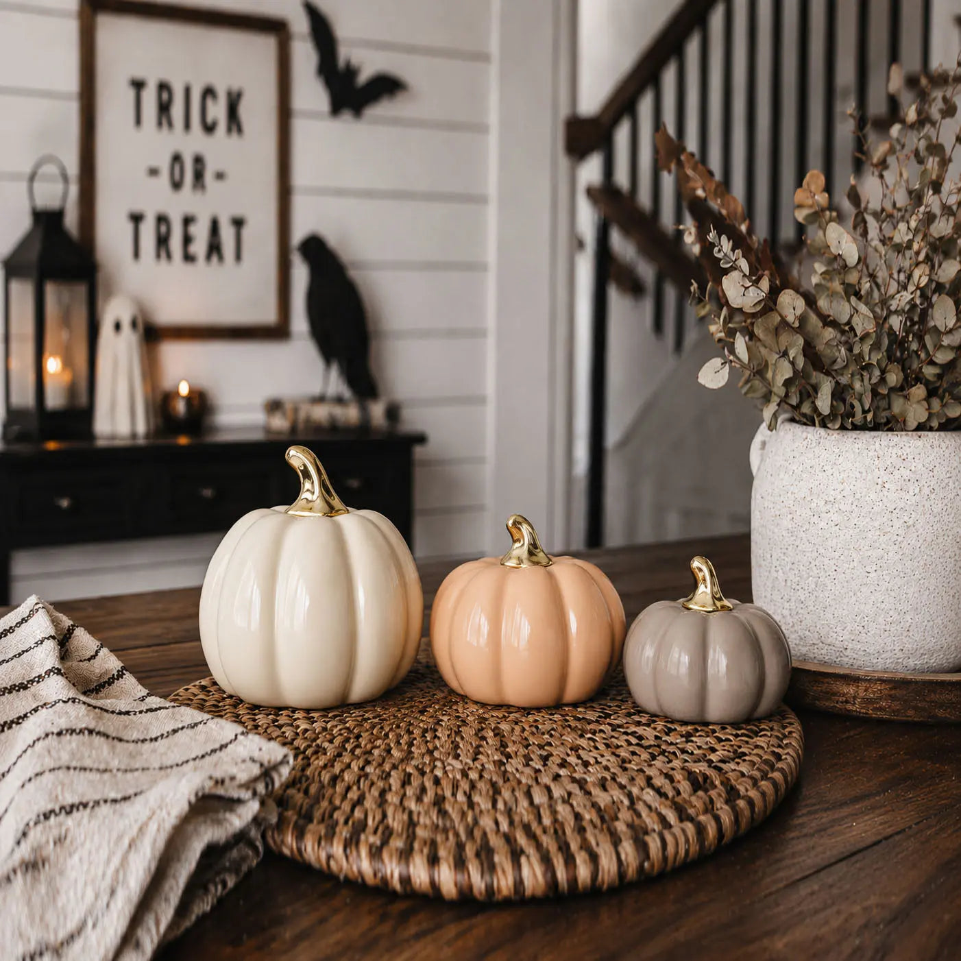 Three small decorative pumpkins in cream, peach, and gray are arranged on a woven round placemat on a wooden table, with a white speckled vase of dried leaves and a framed 'Trick or Treat' sign in the background.