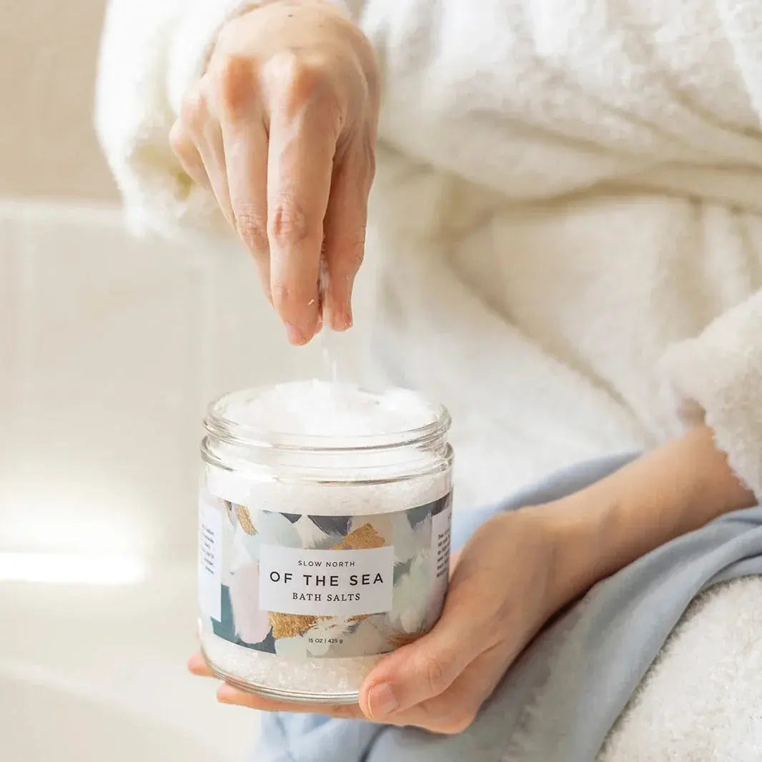 Cozy bath salts in glass jar, labeled 'Of the Sea,' held by person in white robe for relaxing self-care