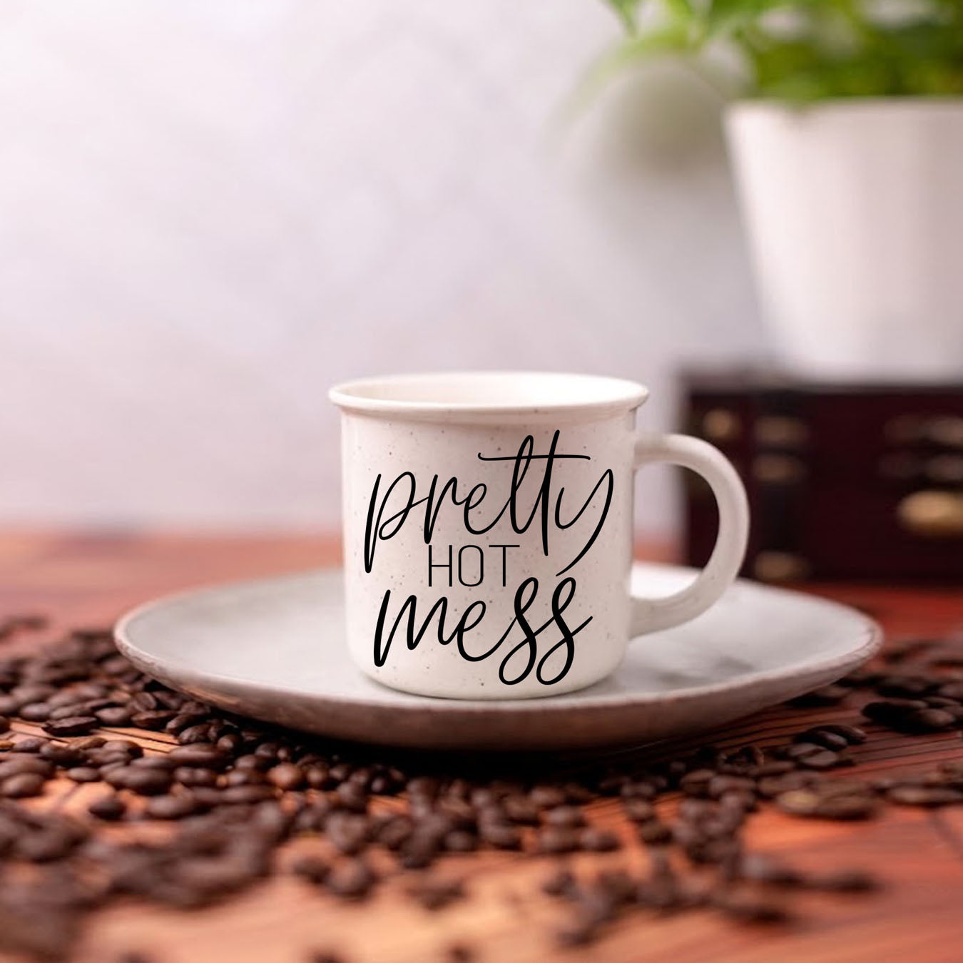 White mug with 'Pretty Hot Mess' text on a saucer surrounded by coffee beans