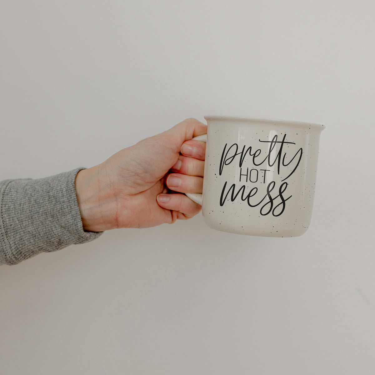 Hand holding a mug with 'pretty hot mess' text against a plain background