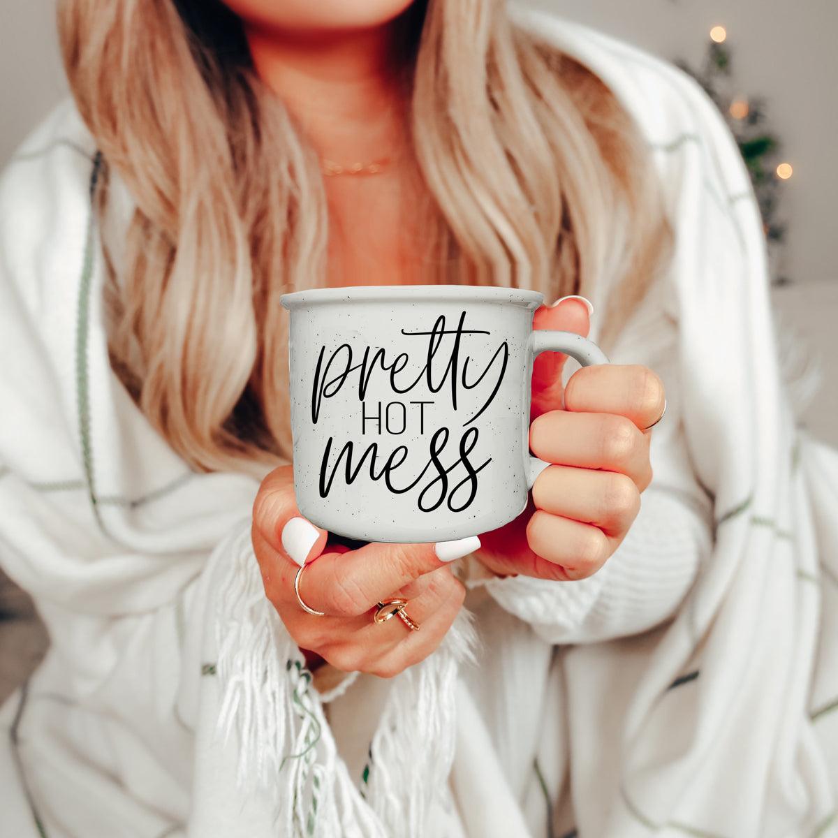 Person holding a mug with 'pretty hot mess' text, wearing a white sweater.
