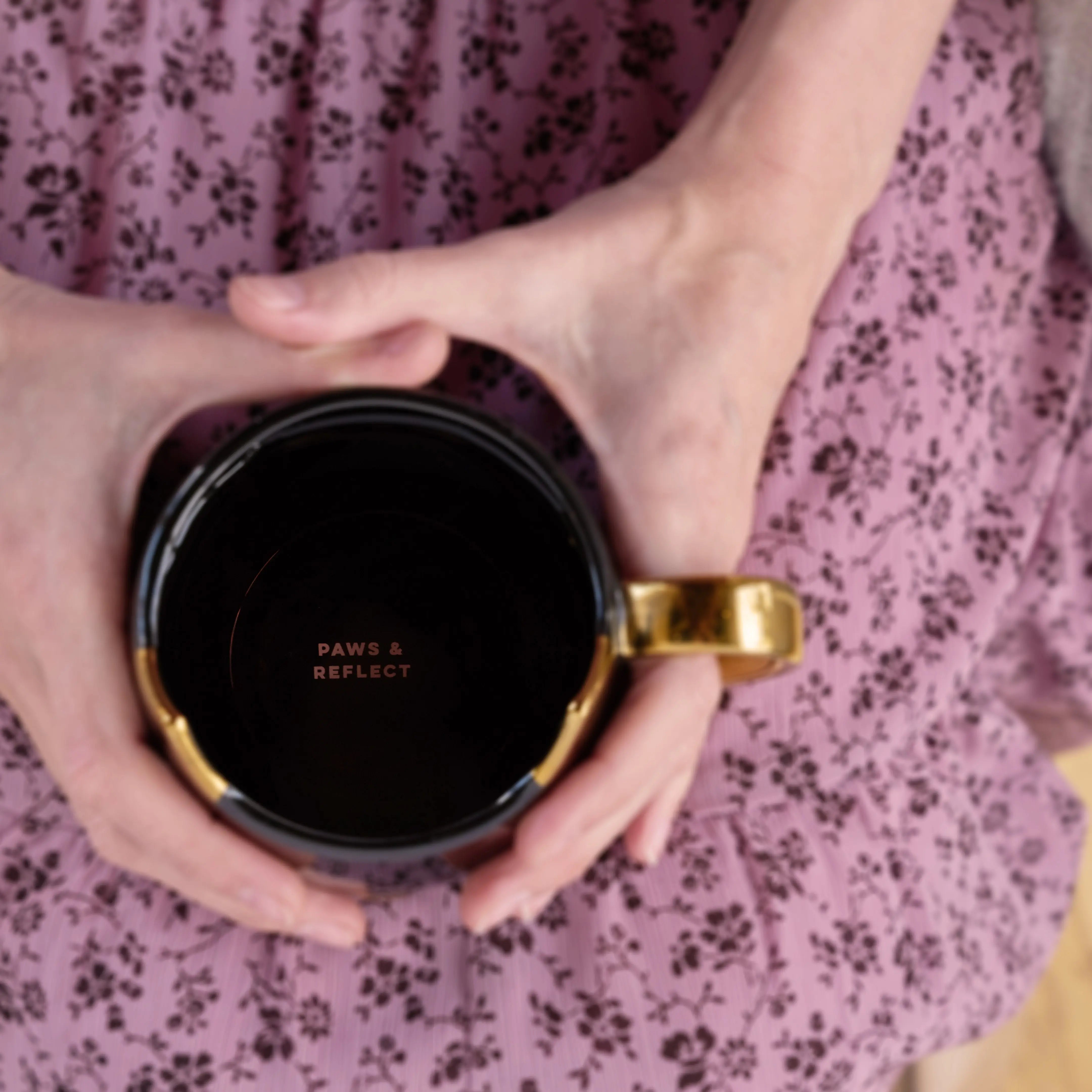 Person holding a black mug with gold handle against a floral-patterned fabric background