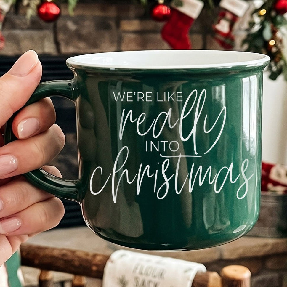 Green mug with 'We're like really into Christmas' text held by a hand, Christmas stockings in the background