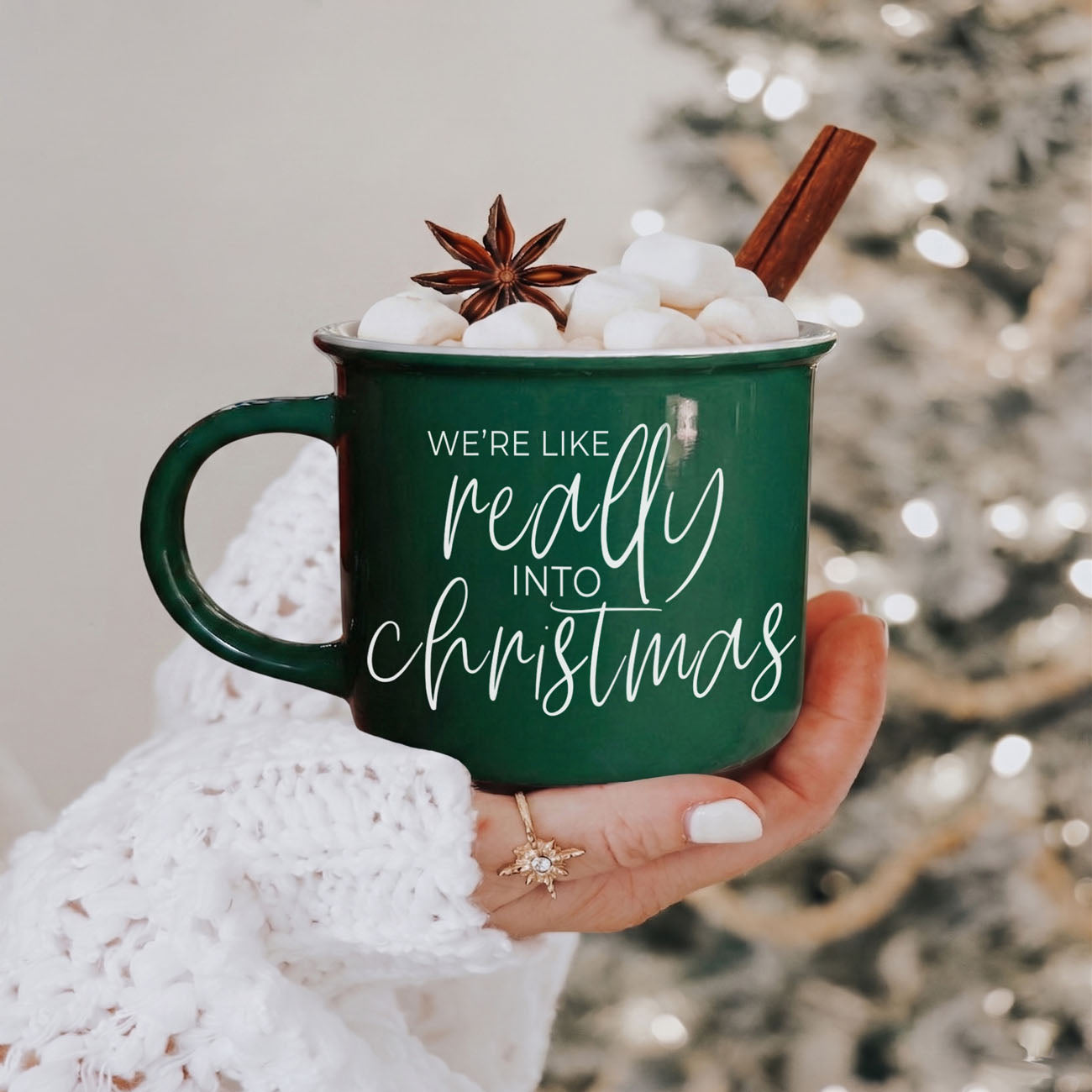 Green mug with 'We're like really into Christmas' text, held by a person, with hot chocolate and garnishes against a blurred festive background.