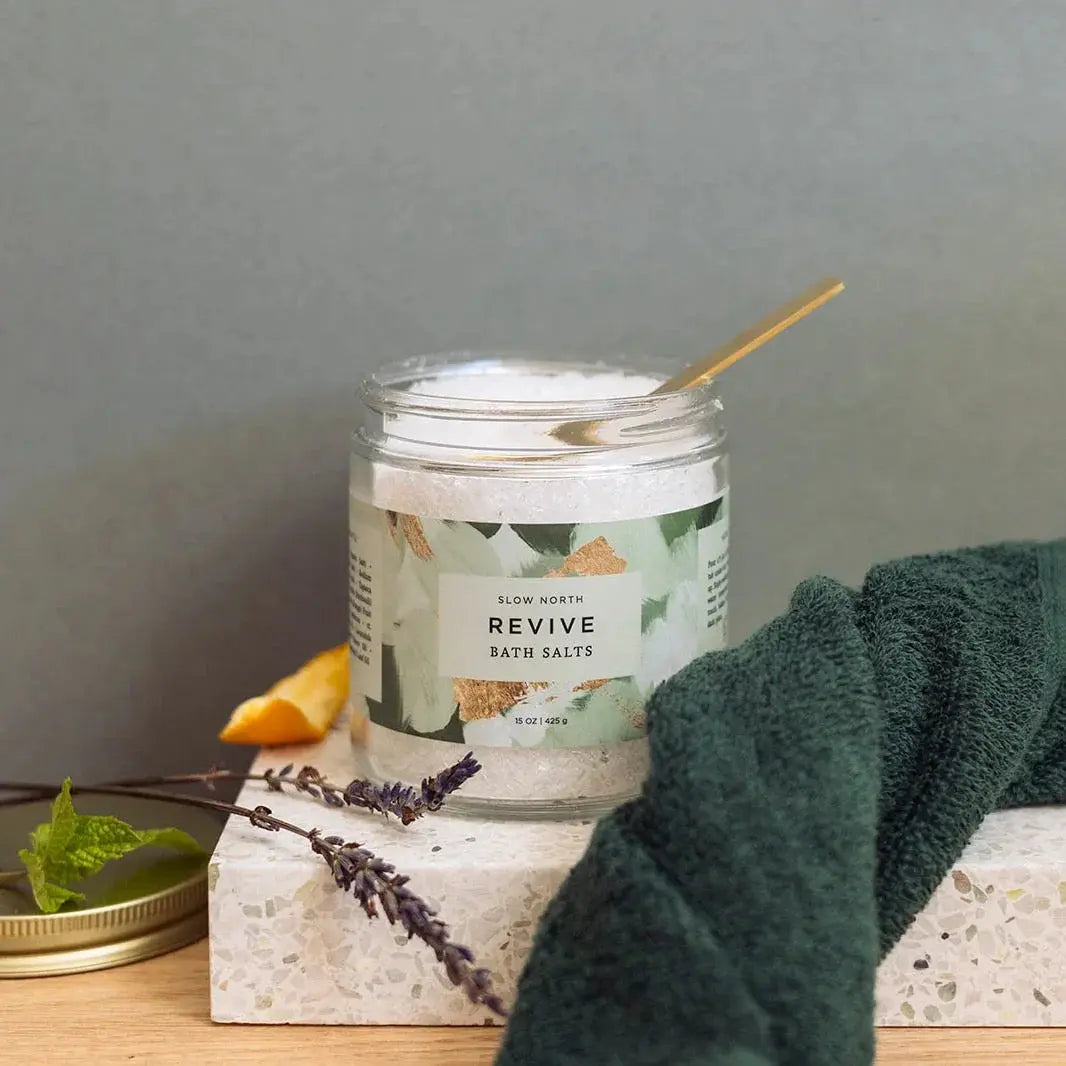 Revive bath salts in glass jar with gold spoon, lavender sprig, and green towel for cozy self-care