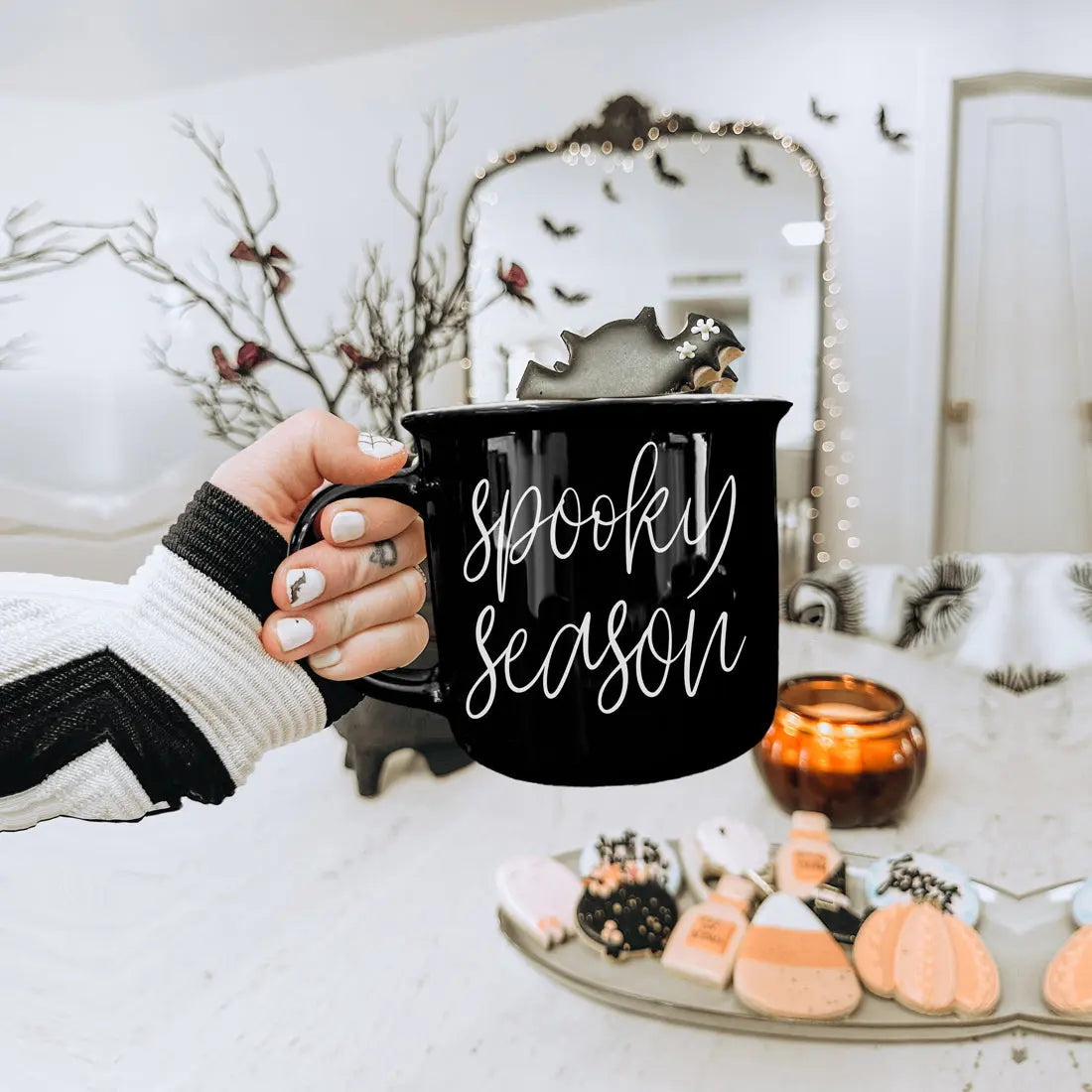 Person holding a black mug with 'Spooky Season' text, surrounded by Halloween-themed decor.