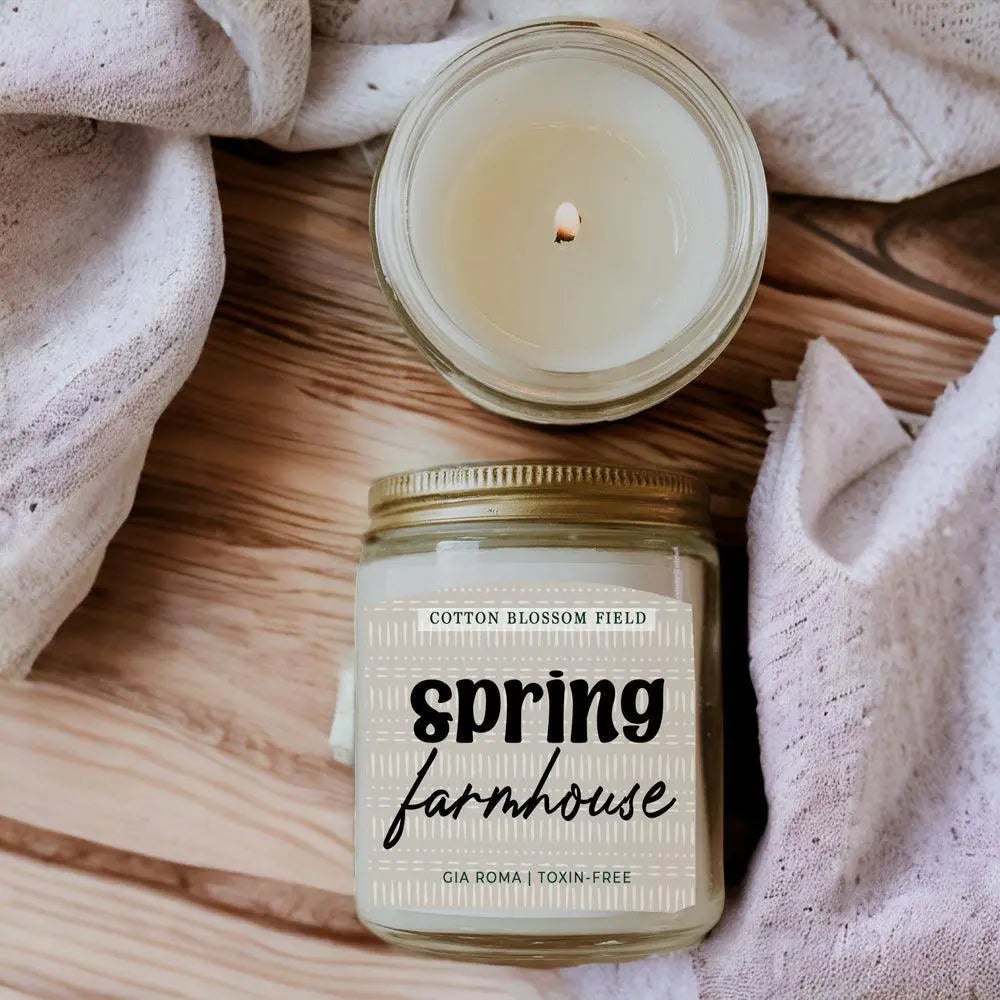 Candle in a jar labeled 'Spring Farmhouse' on a wooden surface with a soft fabric background.