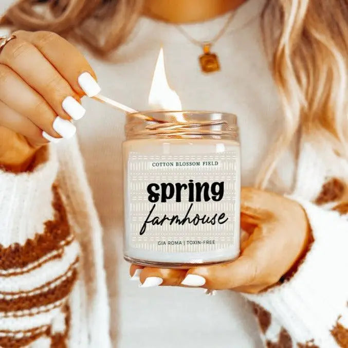 Person lighting a 'Spring Farmhouse' candle with cotton blossom field branding.