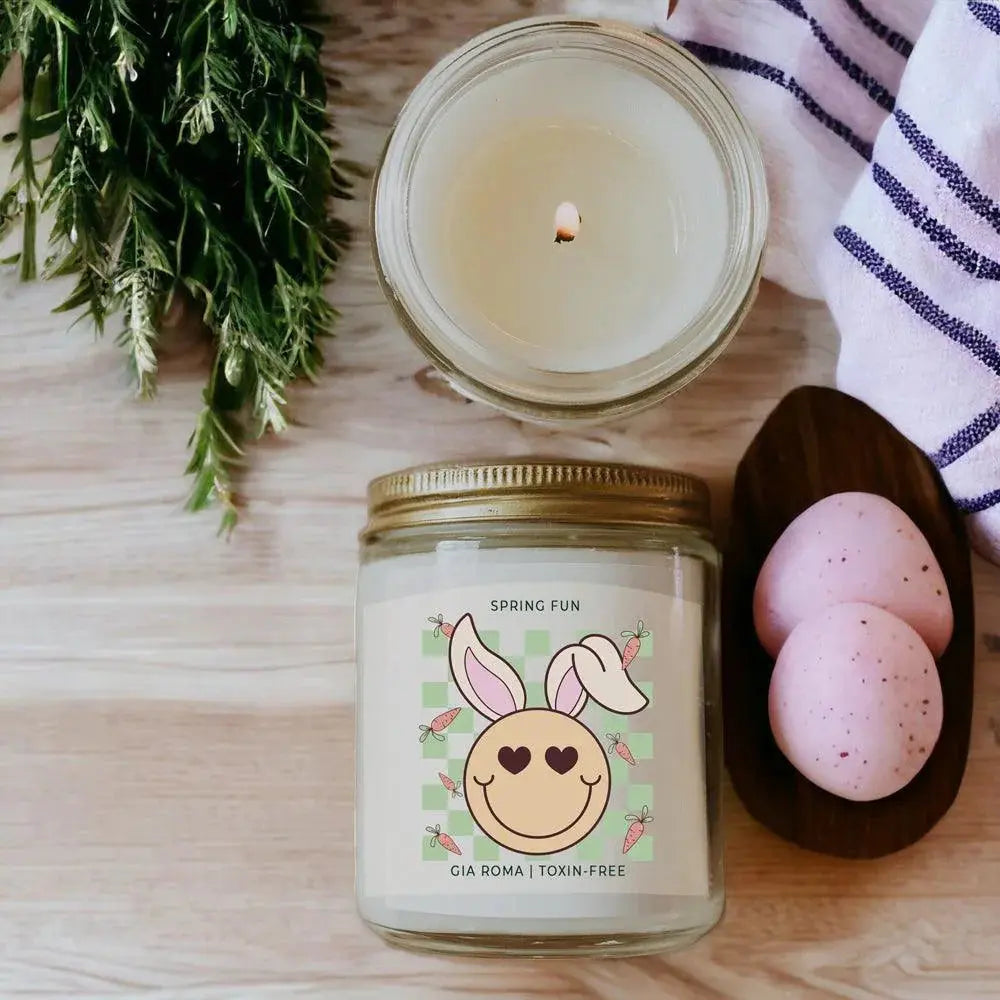 Candle jar with a decorative label featuring a bunny face, next to pink Easter eggs and greenery on a wooden surface.