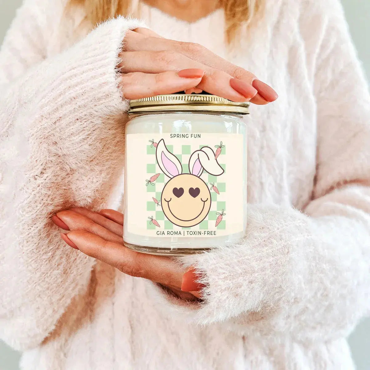 Person holding a candle jar with a bunny design label, wearing a fluffy white sweater.