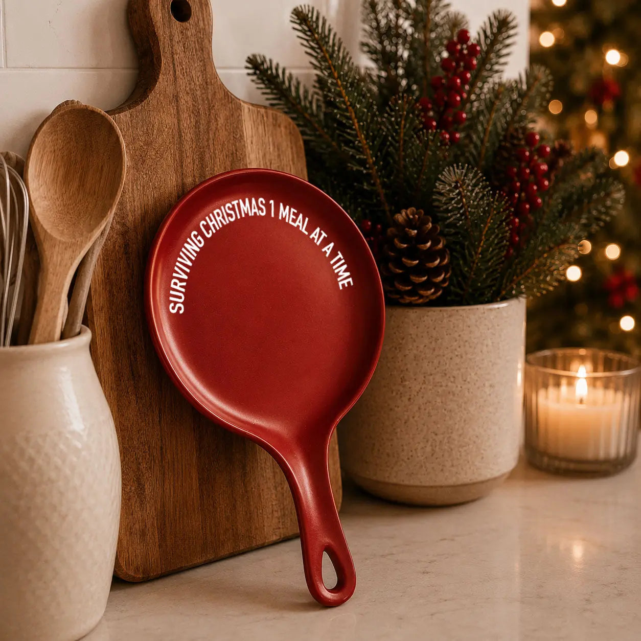Red mini cast iron skillet with the phrase "Surviving Christmas 1 Meal at a Time" displayed against a wooden cutting board beside wooden kitchen utensils, fresh evergreen branches, red berries, pinecones, and a glowing candle, creating a cozy farmhouse Christmas kitchen display.