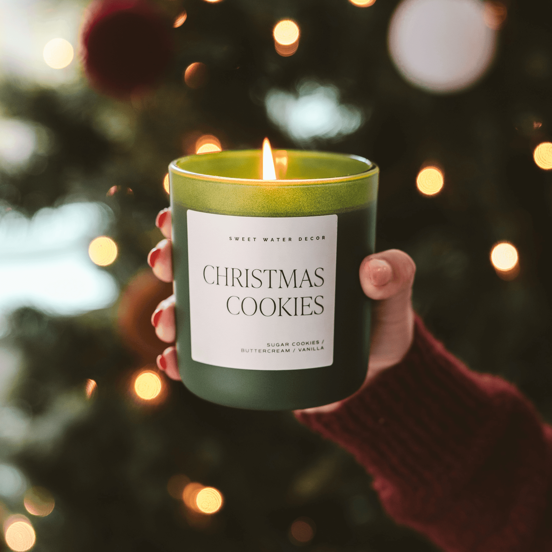 Hand holding a 'Christmas Cookies' candle with festive lights and decorations in the background