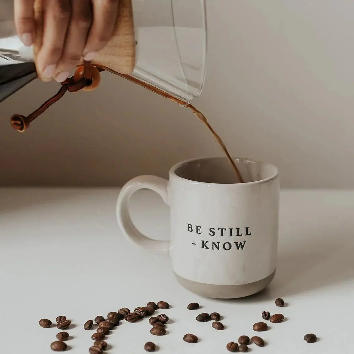 White ceramic mug with 'Be Still + Know' text, coffee being poured, scattered coffee beans, cozy farmhouse decor