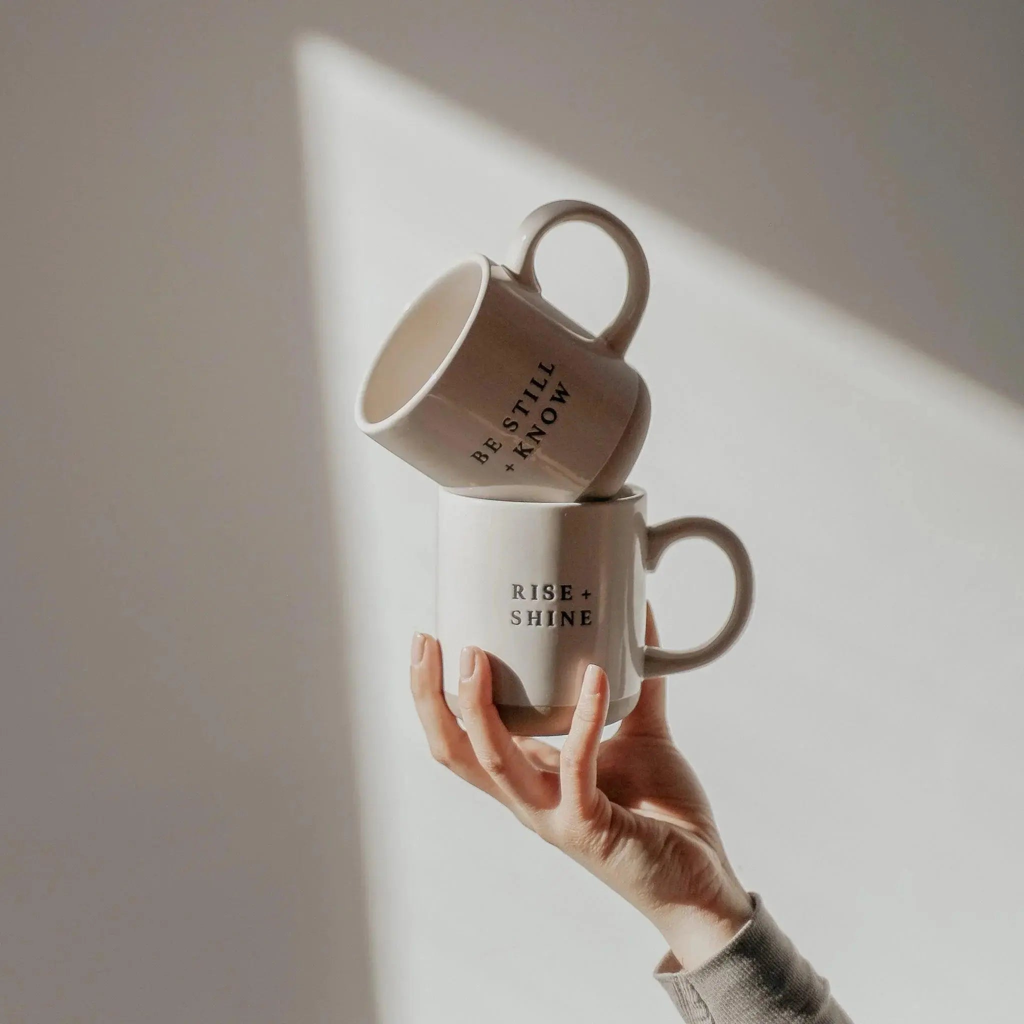 Hand holding two stacked ceramic mugs with 'Rise + Shine' and 'Be Still + Know' quotes, cozy farmhouse decor