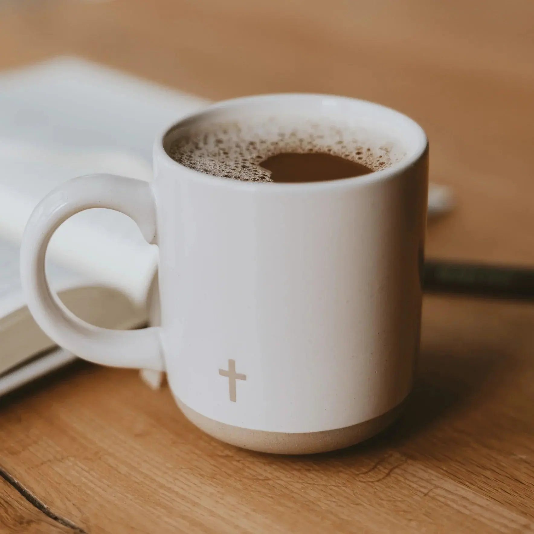 White ceramic coffee mug with cross design, cozy farmhouse style, on wooden table beside book