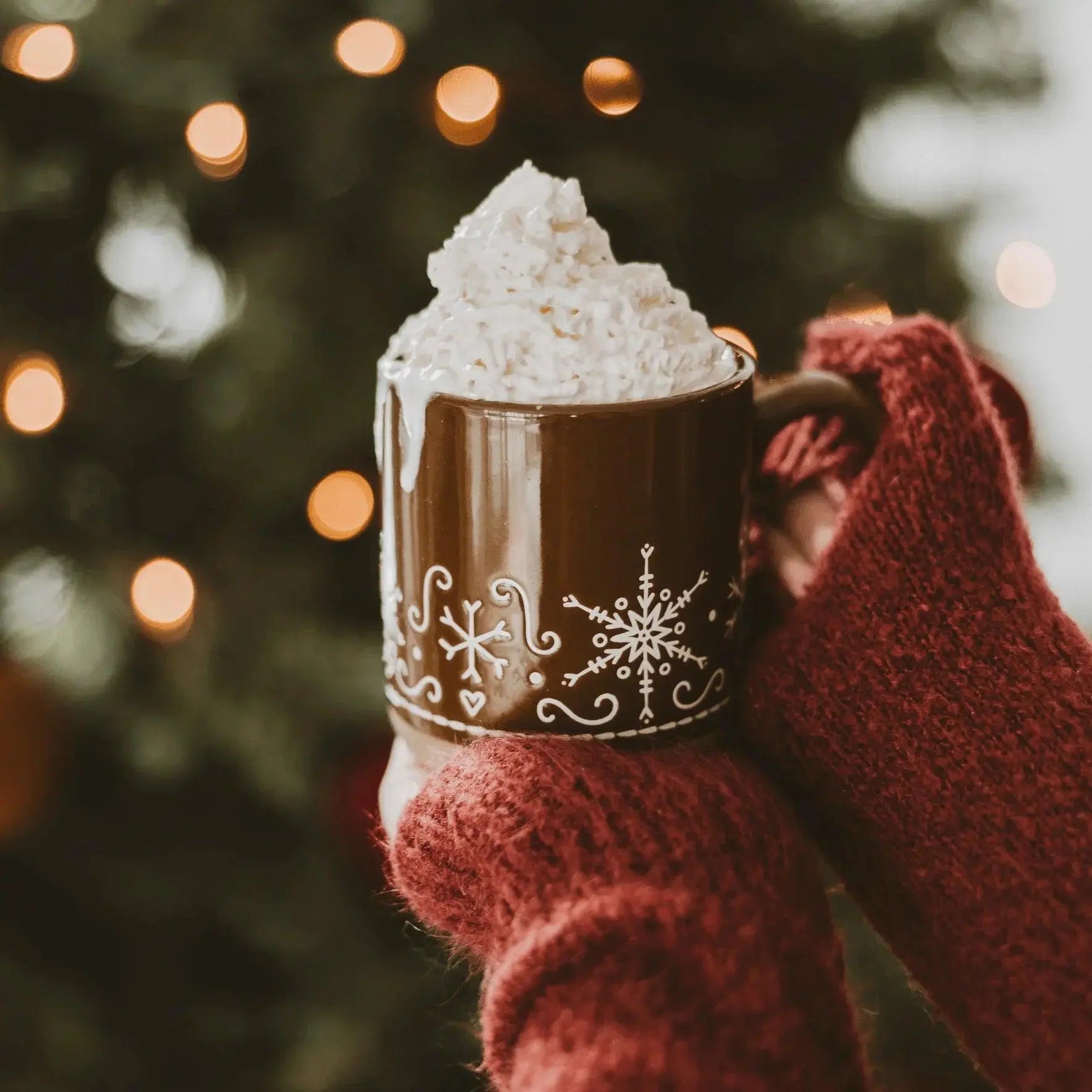 Cozy winter mug with snowflake design, topped with whipped cream, held in red mittens near festive lights