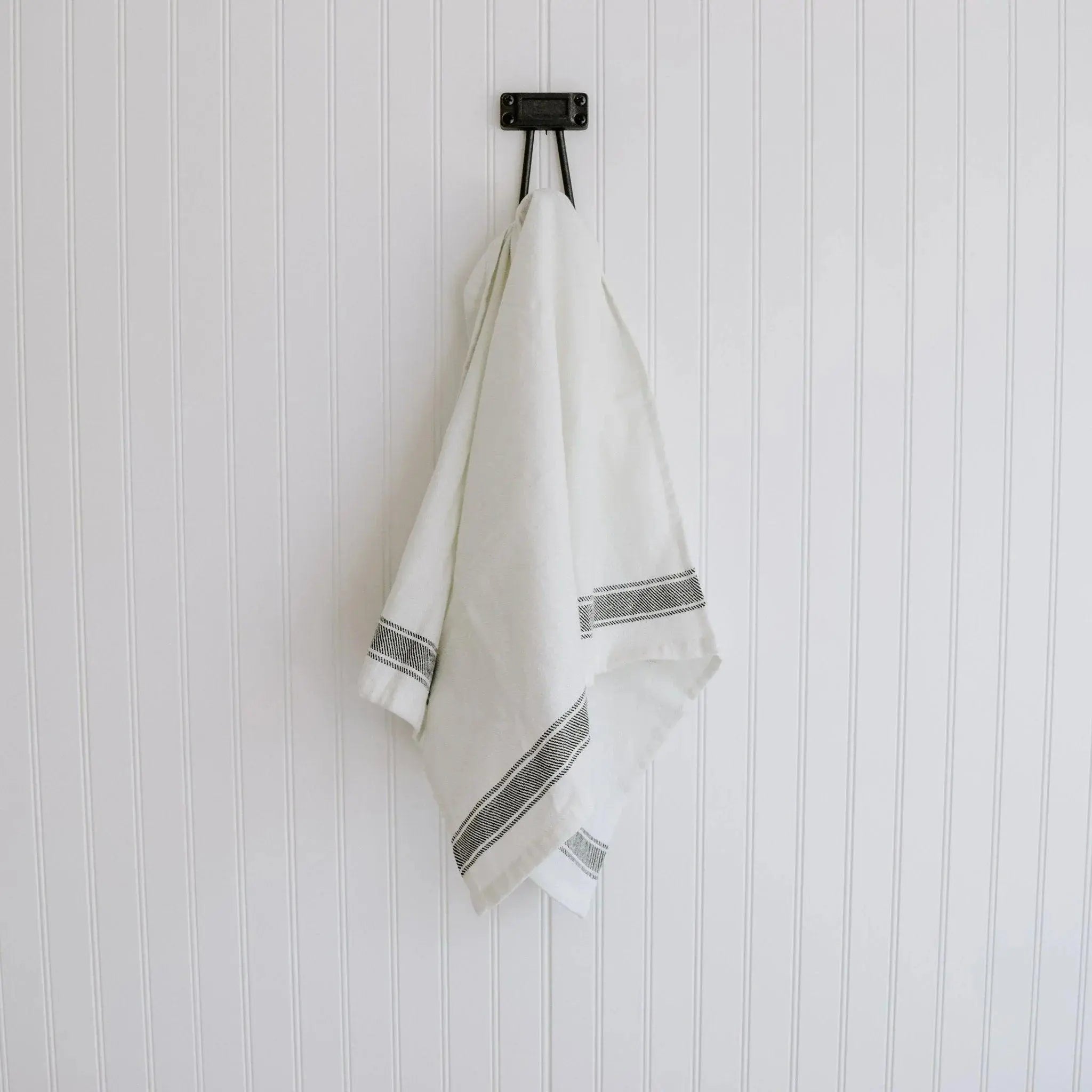 White farmhouse kitchen towel with black stripes hanging on rustic hook against beadboard wall