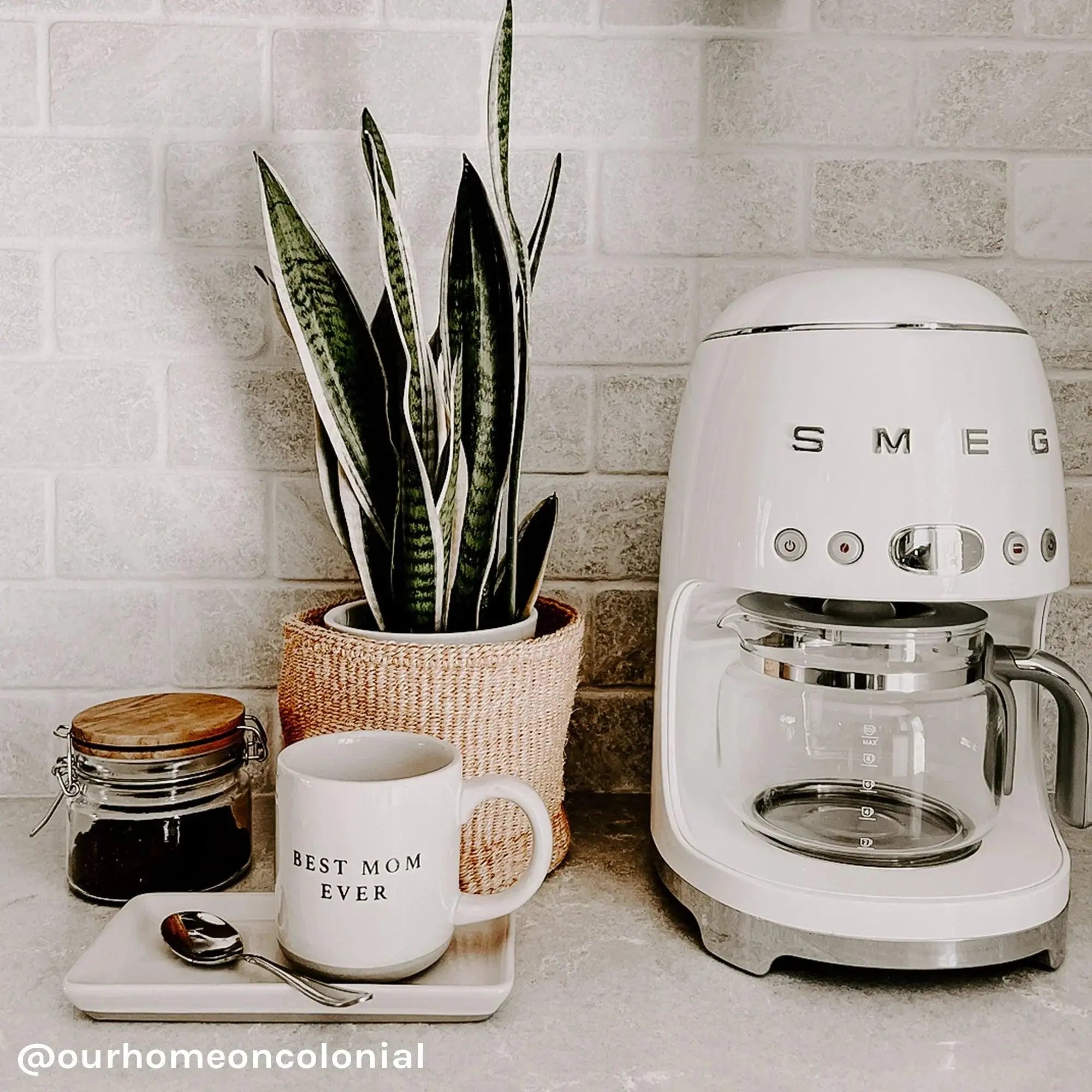 White SMEG coffee maker with snake plant, 'Best Mom Ever' mug, and coffee jar on cozy kitchen counter