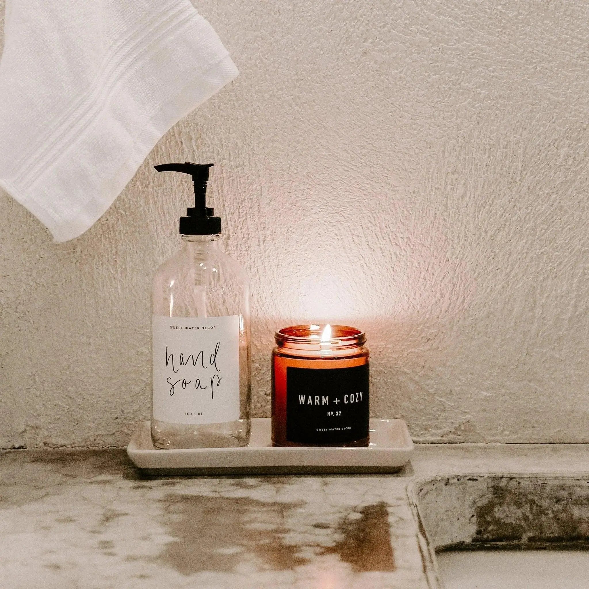 Cozy bathroom setup with hand soap dispenser and warm + cozy candle on marble countertop