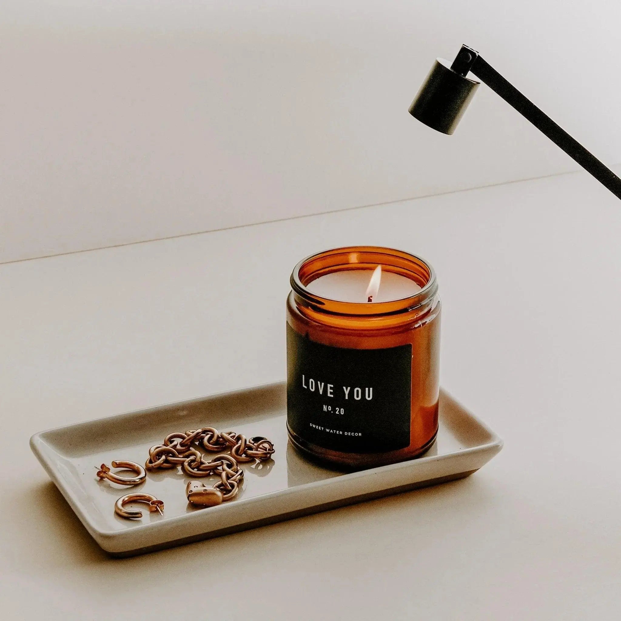 Lit amber candle labeled 'Love You No. 20' on ceramic tray with gold jewelry, modern lamp nearby