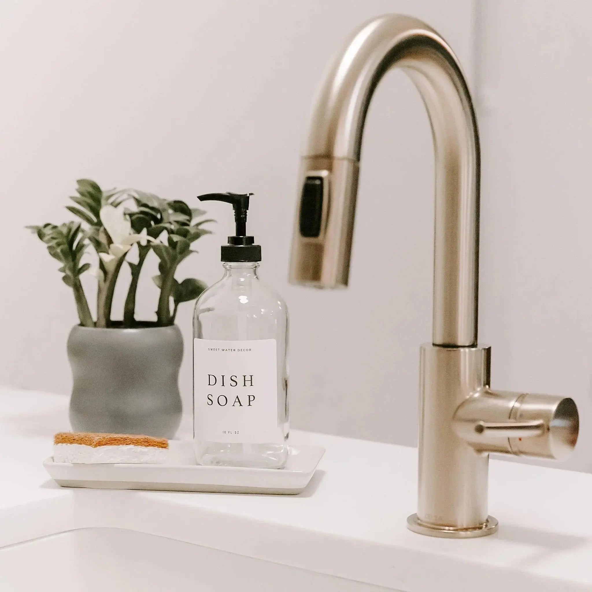 Glass dish soap bottle with black pump, small potted plant, and sponge on white tray beside modern kitchen faucet