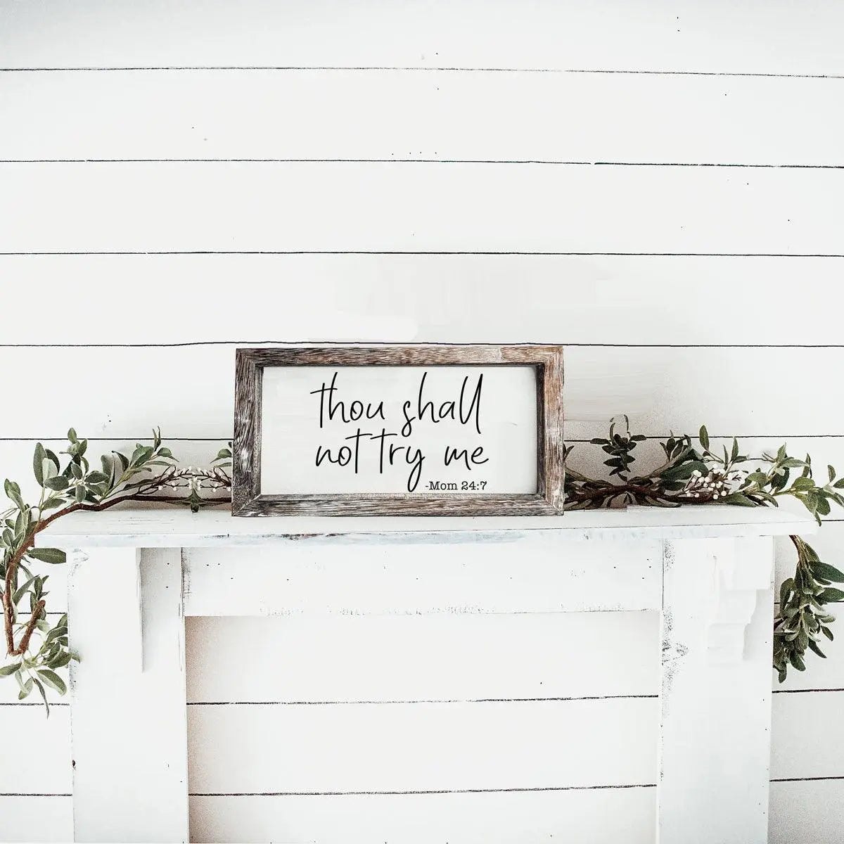 Framed sign with 'thou shall not try me' on a white wooden wall with greenery.