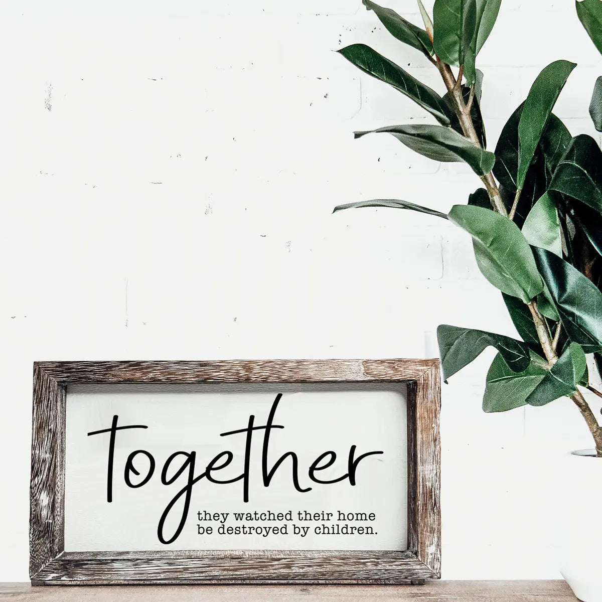Framed artwork with the word 'Together' on a white wall with a plant.