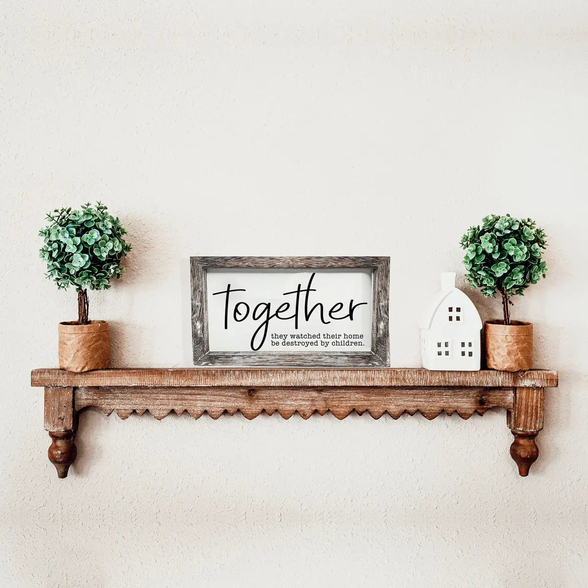 Wooden shelf with decorative items including a framed sign, potted plants, and a small house-shaped object on a light beige wall.