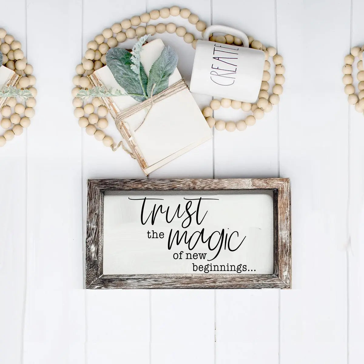 Decorative setup with a framed sign, mug, notebook, and plant on a white wooden surface.
