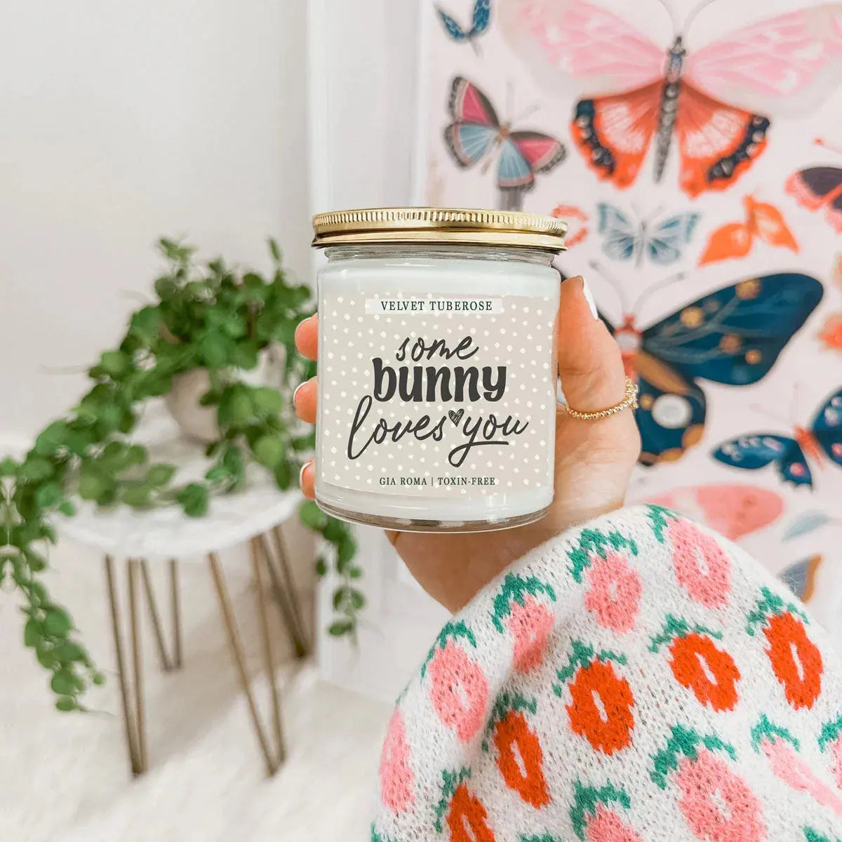 Person holding a candle jar labeled 'some bunny loves you' in front of a colorful butterfly-patterned fabric.
