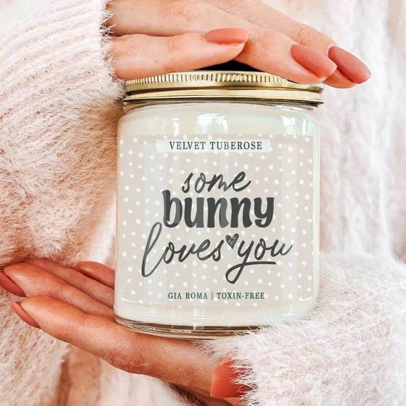 Candle jar labeled 'some bunny loves you' held by a person against a soft pink background