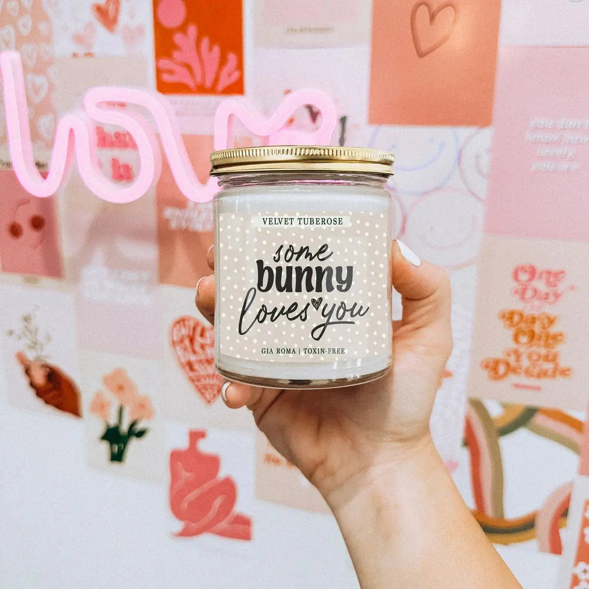 Hand holding a candle jar labeled 'some bunny loves you' against a decorative background with hearts and text.