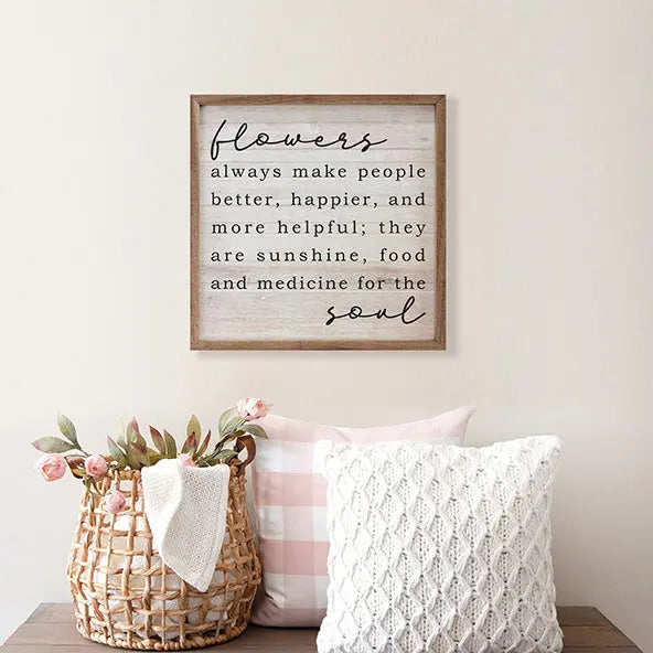 Decorative wall sign with floral quote, basket with flowers, and pillows on a wooden surface.