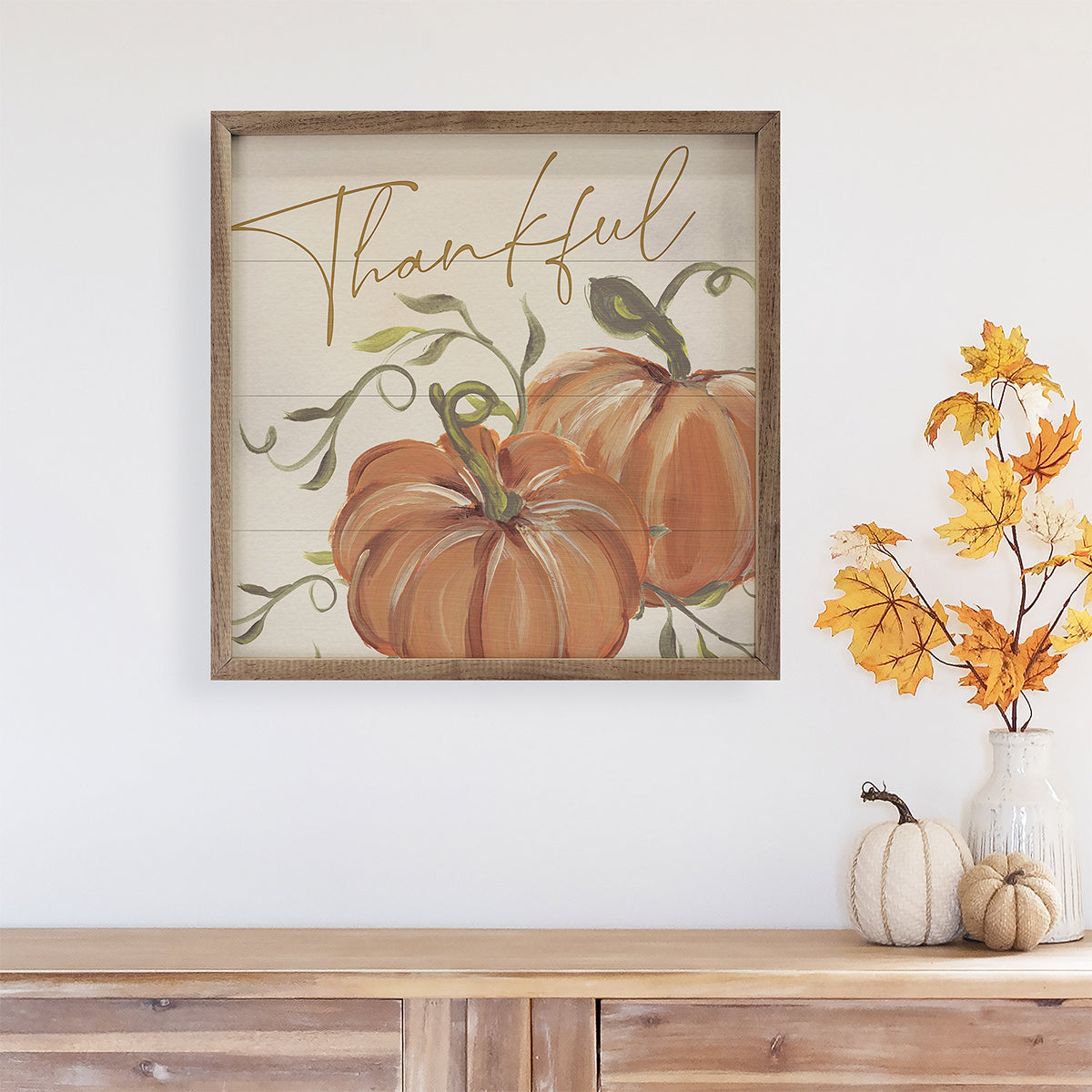 Framed artwork with pumpkins and 'Thankful' text above a wooden surface with decorative items.
