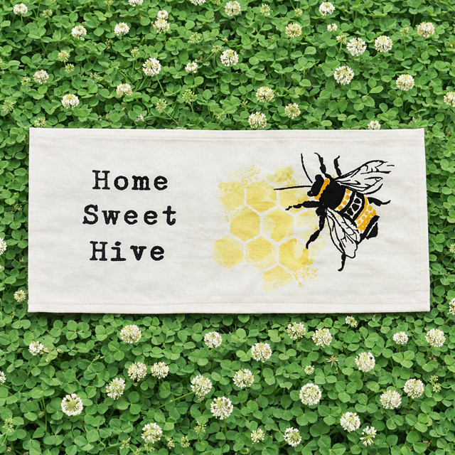 A rectangular white fabric sign or towel laid flat on a bed of green clover with small white flowers. The fabric has black text reading 'Home Sweet Hive' and a yellow honeycomb pattern with a black-and-yellow bumblebee illustration.