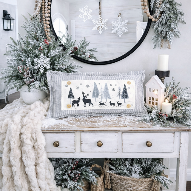 Decorative Christmas setting with a pillow featuring reindeer, trees, and snowflakes on a white dresser.