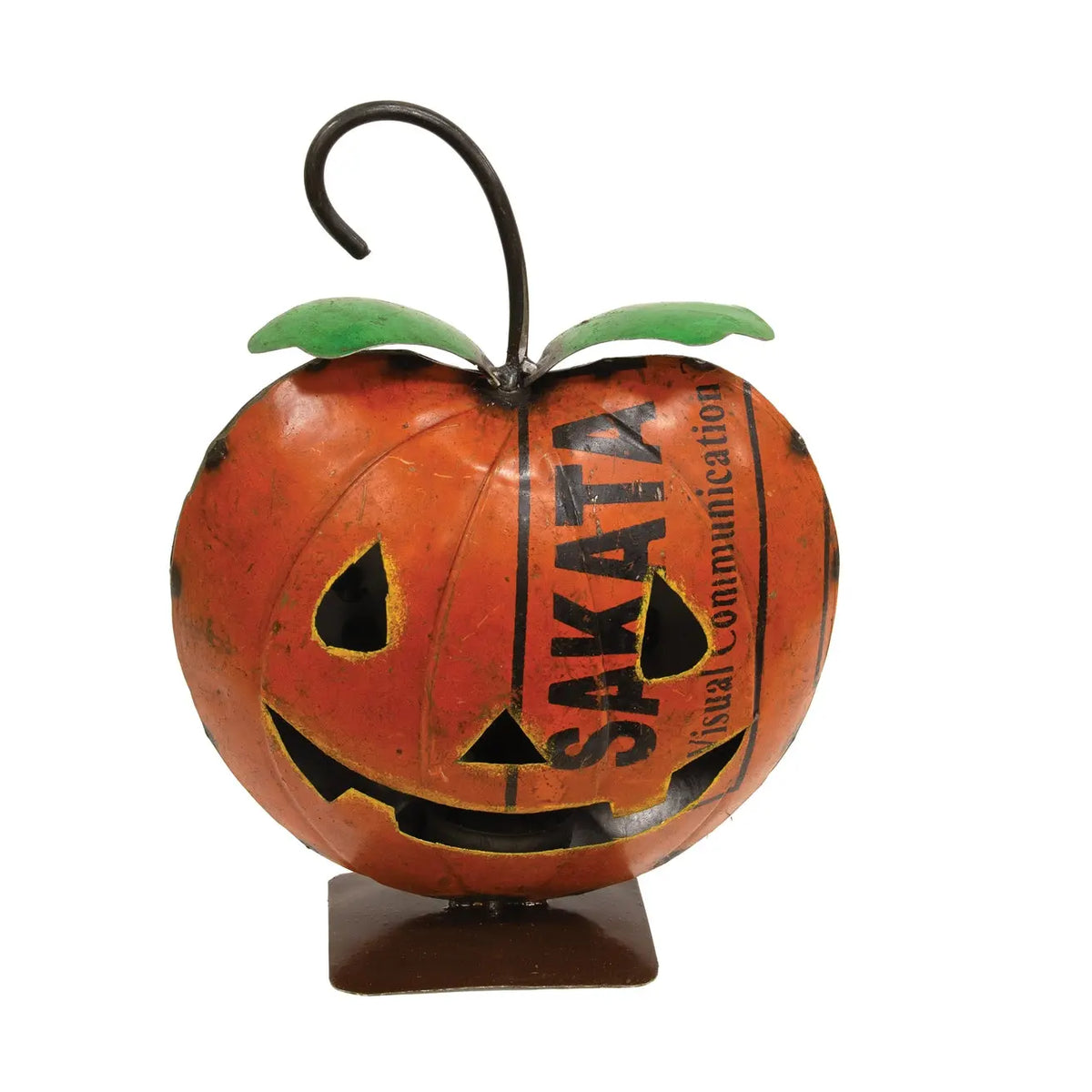 A decorative metal pumpkin sculpture with a carved jack-o'-lantern face, green leaves, and a hook handle on a stand. The front is printed with the text 'SAKATA Visual Communication'.