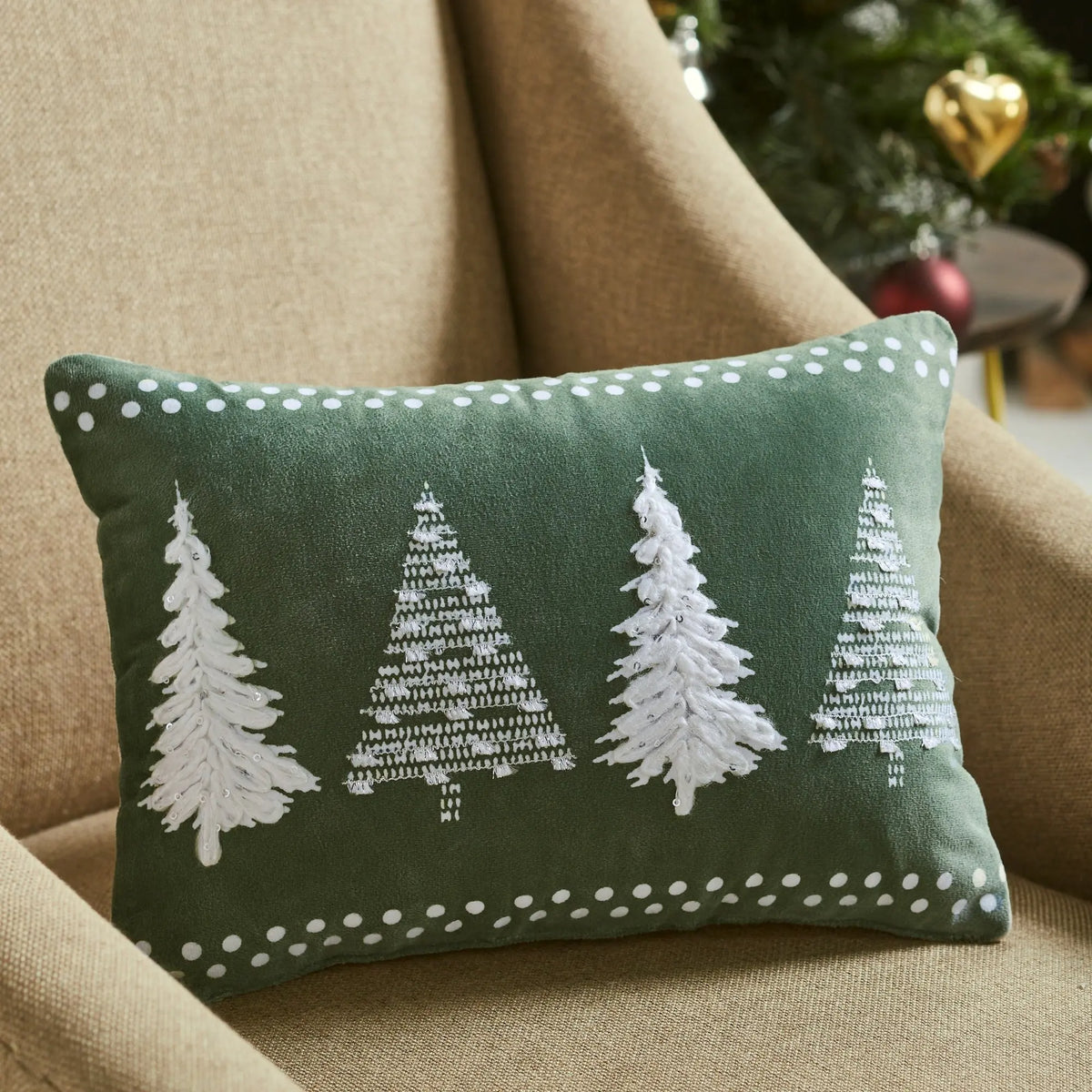 A rectangular green throw pillow with four white embroidered Christmas trees and white dotted borders, resting on a beige armchair with a blurred holiday tree in the background.