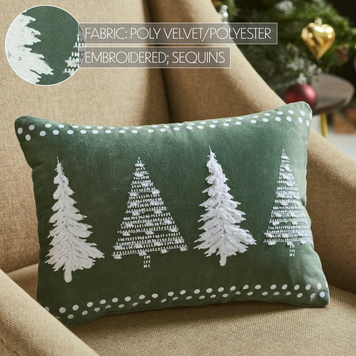 A rectangular green throw pillow with four white embroidered Christmas trees and white polka dot trim, resting on a beige armchair. A circular fabric detail inset is in the top-left corner, and a blurred holiday background is visible.